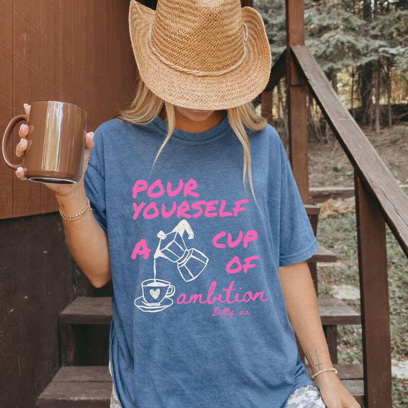 Pu&ograve; includere: Una t-shirt blu con la grafica di una moka che versa caff&egrave; in una tazza e il testo "POUR YOURSELF A CUP OF AMBITION" in rosa e bianco. La maglietta &egrave; abbinata a un cappello da cowboy in paglia e a una tazza in ceramica marrone.