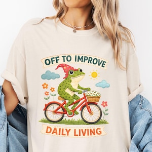 May include: A beige t-shirt featuring a graphic of a green frog wearing a red hat with yellow stars, riding a red bicycle with a basket of white flowers. The text reads OFF TO IMPROVE DAILY LIVING, surrounded by blue clouds, a sun, and orange flowers.
