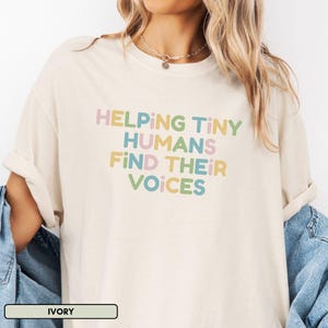 Peut inclure: Un t-shirt beige clair avec le texte "Helping Tiny Humans Find Their Voices" en lettres colorées.