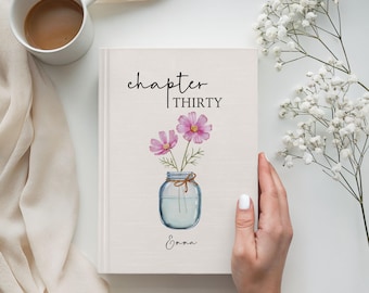 Może przedstawiać: Twarda oprawa dziennika z napisem "chapter THIRTY" i imieniem "Emma" na okładce. Okładka przedstawia akwarelową ilustrację różowych kwiatów w szklanym słoiku. W pobliżu znajduje się biały kubek kawy.