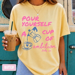 Pu&ograve; includere: Una t-shirt a maniche corte color giallo burro con la grafica di una moka che versa caff&egrave; in una tazzina con un cuore. Il testo recita: POUR YOURSELF A CUP OF ambition Dolly xo. La maglietta presenta un girocollo e una vestibilit&agrave; ampia.