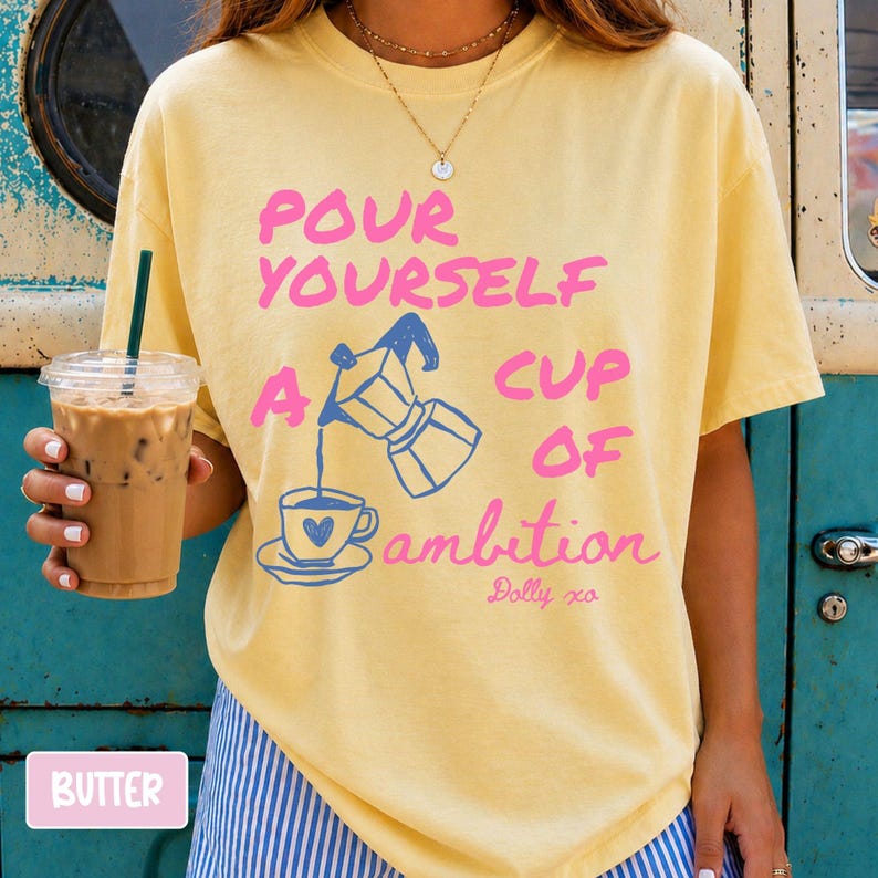 Pu&ograve; includere: Una t-shirt a maniche corte color giallo burro con la grafica di una moka che versa caff&egrave; in una tazzina con un cuore. Il testo recita: POUR YOURSELF A CUP OF ambition Dolly xo. La maglietta presenta un girocollo e una vestibilit&agrave; ampia.