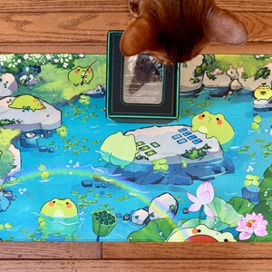 Puede incluir: Una colorida alfombrilla de juego con una escena de estanque caprichosa con ranas de dibujos animados, nen&uacute;fares y flores de loto. La alfombrilla tiene un esquema de color azul y verde con un arco&iacute;ris. Un tarjetero negro est&aacute; encima.