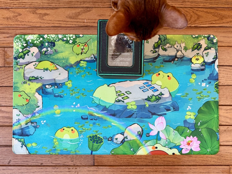 Puede incluir: Una colorida alfombrilla de juego con una escena de estanque caprichosa con ranas de dibujos animados, nen&uacute;fares y flores de loto. La alfombrilla tiene un esquema de color azul y verde con un arco&iacute;ris. Un tarjetero negro est&aacute; encima.