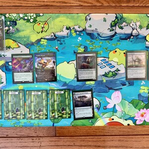 Puede incluir: Una colorida alfombrilla de juego con un dise&ntilde;o de estanque con ranas de dibujos animados y nen&uacute;fares. Varias cartas coleccionables de Magic: The Gathering se muestran en la alfombrilla, sobre una superficie de madera.