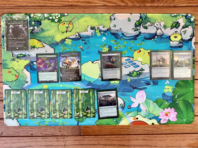 Puede incluir: Una colorida alfombrilla de juego con un dise&ntilde;o de estanque con ranas de dibujos animados y nen&uacute;fares. Varias cartas coleccionables de Magic: The Gathering se muestran en la alfombrilla, sobre una superficie de madera.