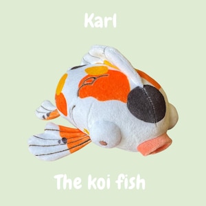 Könnte beinhalten: Ein Plüsch-Koi-Fisch-Spielzeug in Weiß, Orange, Gelb und Schwarz. Das Spielzeug hat ein lächelndes Gesicht und den Text "Karl" und "The koi fish" in Weiß. Der Fisch liegt auf einem hellgrünen Hintergrund.