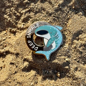 Puede incluir: Un pin de esmalte turquesa y negro con un diseño de tiburón ballena. El pin tiene las palabras "DEEP INHALE" en letras blancas alrededor del borde. El pin está sobre una superficie arenosa.