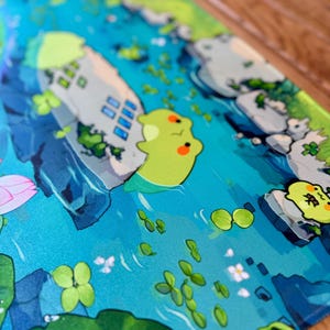 Puede incluir: Alfombrilla de rat&oacute;n ilustrada con una escena acu&aacute;tica caprichosa. El dise&ntilde;o incluye un r&iacute;o azul, edificios de estilo caricatura, nen&uacute;fares y adorables criaturas verdes con mejillas naranjas. La est&eacute;tica general es brillante y juguetona.