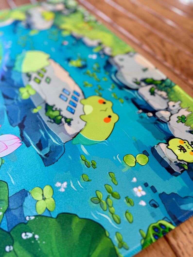 Puede incluir: Alfombrilla de rat&oacute;n ilustrada con una escena acu&aacute;tica caprichosa. El dise&ntilde;o incluye un r&iacute;o azul, edificios de estilo caricatura, nen&uacute;fares y adorables criaturas verdes con mejillas naranjas. La est&eacute;tica general es brillante y juguetona.
