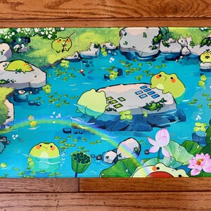 Puede incluir: Alfombrilla de rat&oacute;n rectangular con una ilustraci&oacute;n caprichosa de una escena de estanque con ranas de dibujos animados. El dise&ntilde;o incluye nen&uacute;fares, rocas, un arco&iacute;ris y una flor de loto, todo en colores vibrantes.