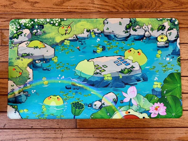 Puede incluir: Alfombrilla de rat&oacute;n rectangular con una ilustraci&oacute;n caprichosa de una escena de estanque con ranas de dibujos animados. El dise&ntilde;o incluye nen&uacute;fares, rocas, un arco&iacute;ris y una flor de loto, todo en colores vibrantes.