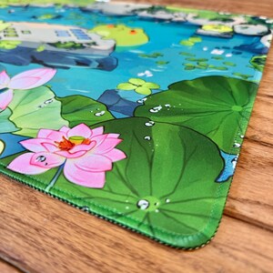 Puede incluir: Alfombrilla de rat&oacute;n colorida con una serena escena de estanque con flores de loto rosas, hojas de nen&uacute;far y una rana. El dise&ntilde;o incluye gotas de agua y una ilustraci&oacute;n detallada de un entorno acu&aacute;tico tranquilo. La alfombrilla tiene un borde verde.