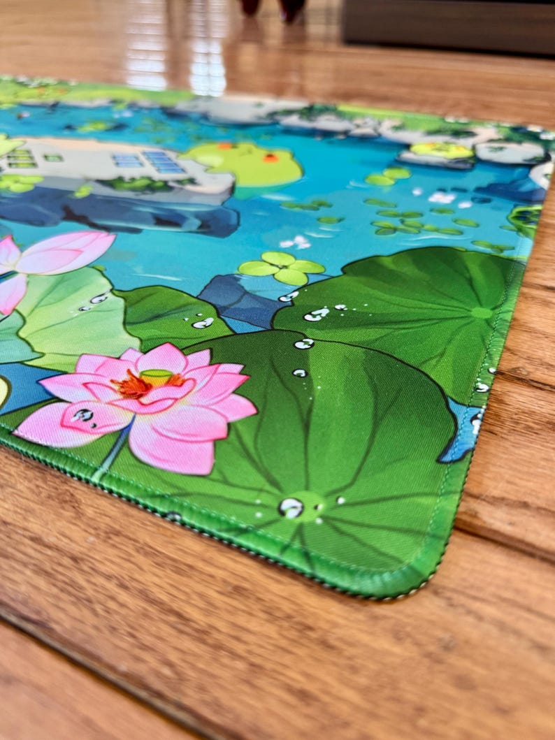 Puede incluir: Alfombrilla de rat&oacute;n colorida con una serena escena de estanque con flores de loto rosas, hojas de nen&uacute;far y una rana. El dise&ntilde;o incluye gotas de agua y una ilustraci&oacute;n detallada de un entorno acu&aacute;tico tranquilo. La alfombrilla tiene un borde verde.