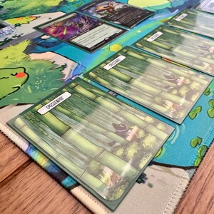 Puede incluir: Una alfombrilla de juego con un dise&ntilde;o acu&aacute;tico colorido, con una rana de dibujos animados y otros elementos acu&aacute;ticos. Se ven varias cartas de juego de cartas coleccionables, incluidas cartas de "Bosque" con ilustraciones de bamb&uacute;.