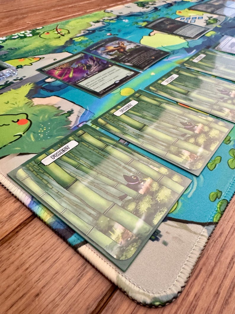 Puede incluir: Una alfombrilla de juego con un dise&ntilde;o acu&aacute;tico colorido, con una rana de dibujos animados y otros elementos acu&aacute;ticos. Se ven varias cartas de juego de cartas coleccionables, incluidas cartas de "Bosque" con ilustraciones de bamb&uacute;.
