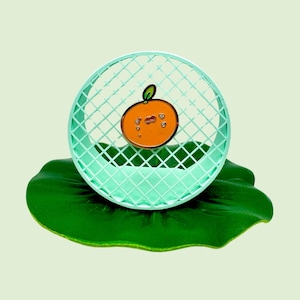 Puede incluir: Un pin de esmalte verde con forma de naranja con una cara sonriente. El pin está sentado sobre una hoja de loto verde.