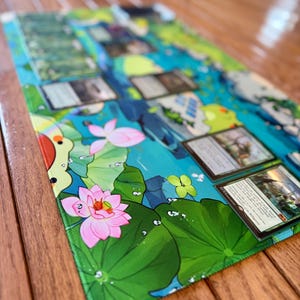 Puede incluir: Una alfombrilla de juego rectangular con una vibrante escena acu&aacute;tica con flores de loto rosas, hojas de nen&uacute;far verdes y un dise&ntilde;o de agua azul y verde. Varias cartas coleccionables est&aacute;n colocadas sobre la alfombrilla, lo que sugiere su uso para juegos de cartas.
