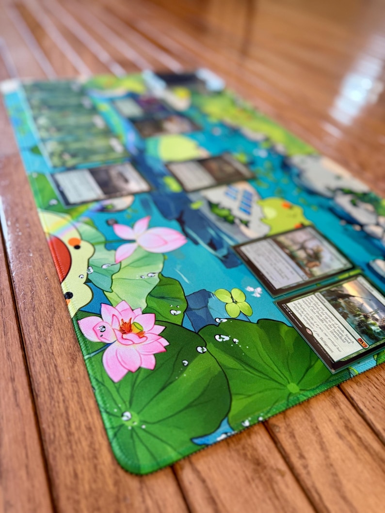 Puede incluir: Una alfombrilla de juego rectangular con una vibrante escena acu&aacute;tica con flores de loto rosas, hojas de nen&uacute;far verdes y un dise&ntilde;o de agua azul y verde. Varias cartas coleccionables est&aacute;n colocadas sobre la alfombrilla, lo que sugiere su uso para juegos de cartas.