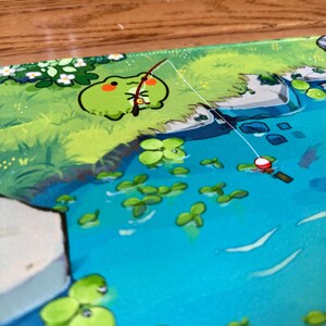 Puede incluir: Alfombrilla de rat&oacute;n caprichosa con una rana de dibujos animados pescando en un estanque. La rana es verde con un rubor rojo, y el estanque es azul con nen&uacute;fares verdes. El fondo incluye follaje verde y flores blancas.