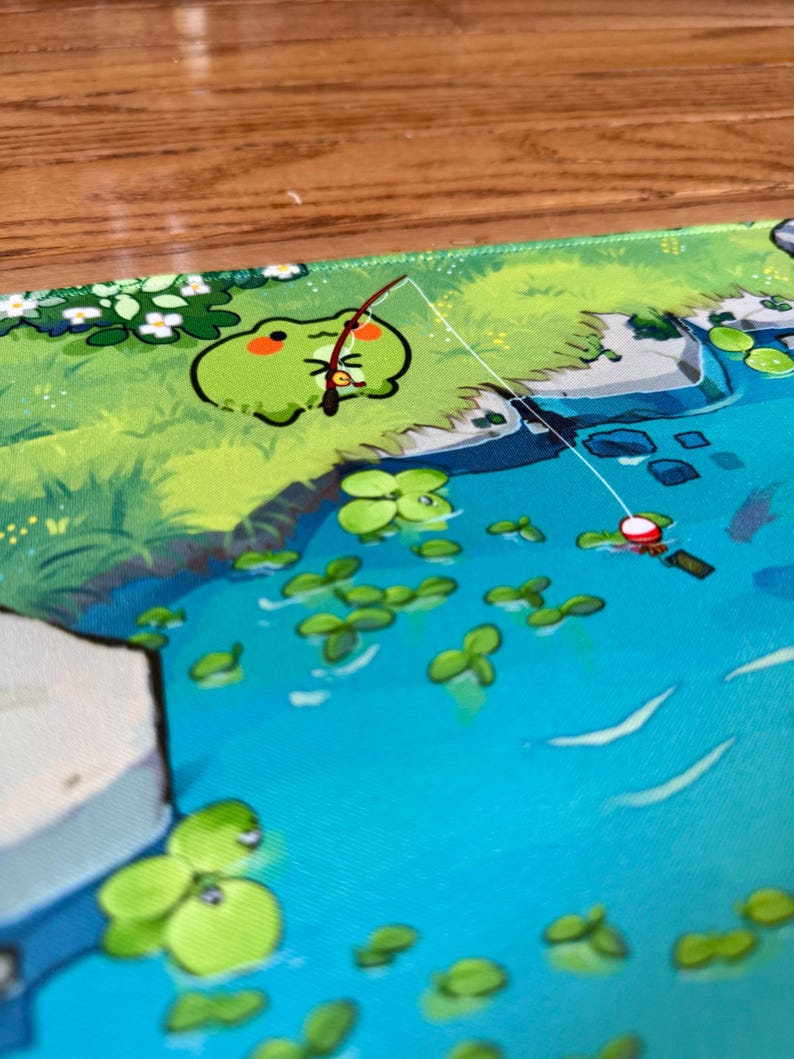 Puede incluir: Alfombrilla de rat&oacute;n caprichosa con una rana de dibujos animados pescando en un estanque. La rana es verde con un rubor rojo, y el estanque es azul con nen&uacute;fares verdes. El fondo incluye follaje verde y flores blancas.