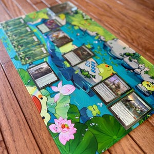 Puede incluir: Una alfombrilla de juego rectangular con un dise&ntilde;o acu&aacute;tico vibrante con flores de loto, hojas de nen&uacute;far y personajes de dibujos animados. Varias cartas coleccionables est&aacute;n dispuestas sobre la alfombrilla. La alfombrilla est&aacute; sobre una superficie de madera.