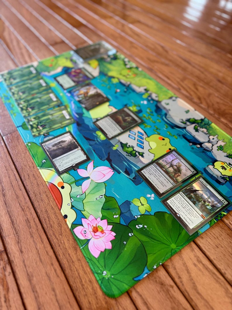 Puede incluir: Una alfombrilla de juego rectangular con un dise&ntilde;o acu&aacute;tico vibrante con flores de loto, hojas de nen&uacute;far y personajes de dibujos animados. Varias cartas coleccionables est&aacute;n dispuestas sobre la alfombrilla. La alfombrilla est&aacute; sobre una superficie de madera.