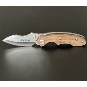 May include: A brown wooden pocket knife with a silver blade. The handle is engraved with the text "RYAN Groom" and the date "18.6.2024".