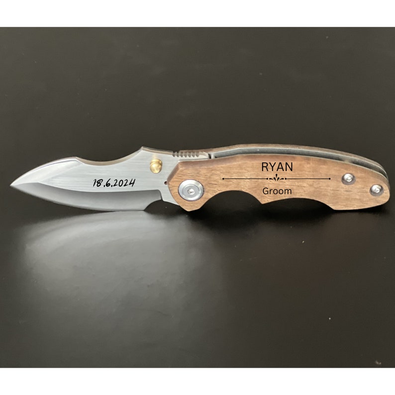 May include: A brown wooden pocket knife with a silver blade. The handle is engraved with the text "RYAN Groom" and the date "18.6.2024".