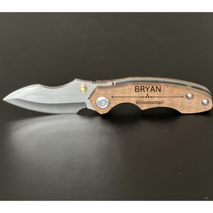 May include: A brown wooden pocket knife with a silver blade. The handle is engraved with the name "Bryan" and the word "Groomsman".