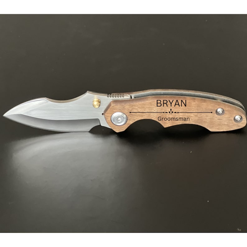 May include: A brown wooden pocket knife with a silver blade. The handle is engraved with the name "Bryan" and the word "Groomsman".
