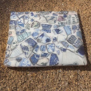 May include: A square coaster with a mosaic design made from broken blue and white ceramic pieces. The fragments are arranged on a white background, creating a unique pattern. The coaster is resting on sand.