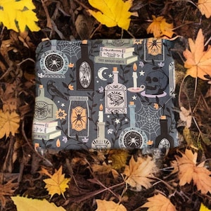 Op de afbeelding: Een zwarte en grijze ritszak met een griezelige Halloween-print. De print bevat verschillende afbeeldingen, waaronder een schedel, spinnen, flessen, kaarsen en boeken. De tekst op de boeken luidt "Age of Alchemy", "Occult Craft" en "500 Ancient Beasts".