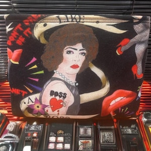 Può includere: Pochette nera con un'illustrazione retrò di una donna con un tatuaggio "BOSS". La borsa presenta un design con tatuaggio a cuore e fiore rosso. La borsa ha anche le parole "DON'T BE IT" e "LIKE".