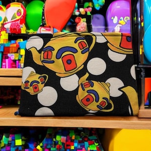 Puede incluir: Bolsa de cosméticos de tela con lunares negros y blancos con un patrón repetido de teteras amarillas con detalles rojos y azules.