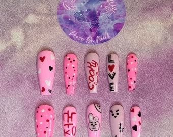 Puede incluir: Conjunto de diez uñas postizas rosas con varios diseños. Algunas uñas tienen lunares negros, corazones y el texto "LOVE". Otras uñas tienen el texto "J-HOPE" y un conejito de dibujos animados.