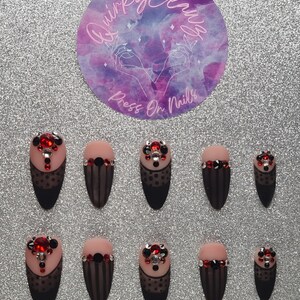 May include: A set of almond-shaped press-on nails with a black and nude color scheme. Each nail features a decorative design with red and clear rhinestones. The nails are arranged on a silver glitter surface, with a circular logo that reads "Quirky Claws Press On Nails."