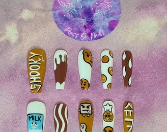May include: A set of ten press-on nails with various designs. The nails feature cartoon characters, cookie and milk themes, and the word "SHOOKY". The nails are in shades of brown, white, and tan, with a glossy finish. A circular logo is in the background.