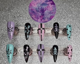 May include: Set of ten press-on nails with various designs. The designs include a pentagram, skulls, a cross with a belt, an infinity symbol with a dagger, and hearts. The nails are in shades of purple, pink, black, and blue.