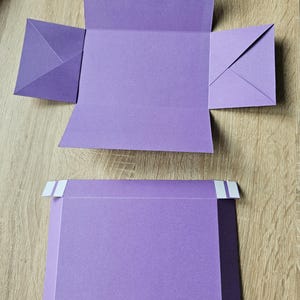 Könnte beinhalten: Zwei Stücke violetter Karton, eines zu einer Schachtelform mit Klappen gefaltet, das andere flach mit weißem Klebeband an den Rändern.