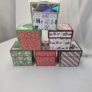 Könnte beinhalten: Ein Stapel dekorativer Weihnachtsgeschenkboxen. Die Boxen weisen verschiedene festliche Muster auf, darunter Zuckerstangen, Lebkuchenmänner und Weihnachtsgrüße. Die Boxen sind in Rot-, Grün- und Weißtönen gehalten, einige haben holografische Akzente.
