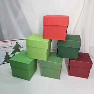 Könnte beinhalten: Sechs farbige, quadratische Geschenkboxen in Rot-, Grün- und Burgundertönen sind pyramidenförmig gestapelt. Im Hintergrund ist eine Weihnachtskarte mit einem Baumdesign zu sehen. Die Boxen sind aus Pappe.