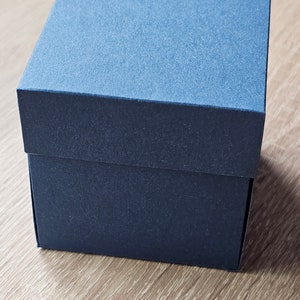 Könnte beinhalten: Eine marineblaue quadratische Geschenkbox mit strukturierter Oberfläche. Die Box ist geschlossen und steht auf einer hellbraunen Holzoberfläche. Die Box hat eine einfarbige Farbe mit passendem Deckel.