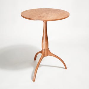 May include: A handcrafted wooden side table with a round top and a tripod base. The table is made of light brown wood and stands approximately 28 inches tall. The table's design is simple and elegant, suitable for various interior styles.