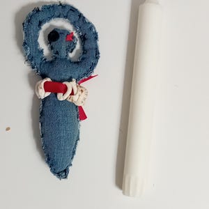 Op de afbeelding: Een denim pop met een spiraalvormige kop, rode lint en schelpaccenten, naast een witte kaars. De pop is gemaakt van blauwe denim stof met gerafelde randen. De kaars is een lange, witte taps toelopende kaars.