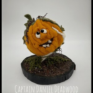 May include: A felt pumpkin with a toothy grin and googly eyes, sitting on a wooden base with moss and dirt. The pumpkin is orange with green vines and a white skeleton arm. The text "Captain Daniel Deadwood" and "PK187" are visible below the pumpkin.