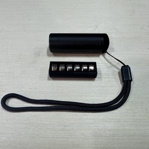 May include: A black cylindrical object with a black wrist strap and a small black rectangular case containing five silver-colored metal objects. The items are arranged on a light-colored surface.
