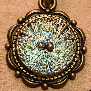 May include: A bronze pendant with a round, iridescent blue and green glass cabochon. The cabochon has a detailed, geometric design with a central cluster of three smaller, copper-colored beads.