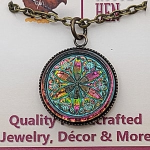 May include: A round iridescent glass pendant with a floral design, hanging from a bronze chain. The pendant has a blue, green, pink, and orange color scheme. The pendant is labeled "Quality handcrafted Jewelry, Décor & More".