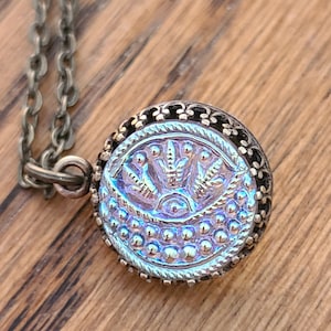 May include: A silver chain necklace with a round pendant featuring an iridescent blue and purple glass button with an ornate design. The button has a raised, stylized eye in the center and a floral pattern around it.